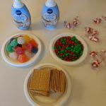 3 small plates arranged in a triangle, one filled with gumdrops, one of m&ms and one of graham crackers. Two bottles of frosting stand above the triangle and peppermint candies are scattered to their right.