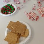 Graham crackers and red and green m&ms on two separate plates surrounded by warped peppermints and smarties