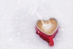 Heart shaped mug filled with steaming coffee sitting in the snow.