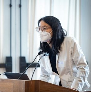 A young woman in a lab coat and mask leans forward to address her audience from across the podium