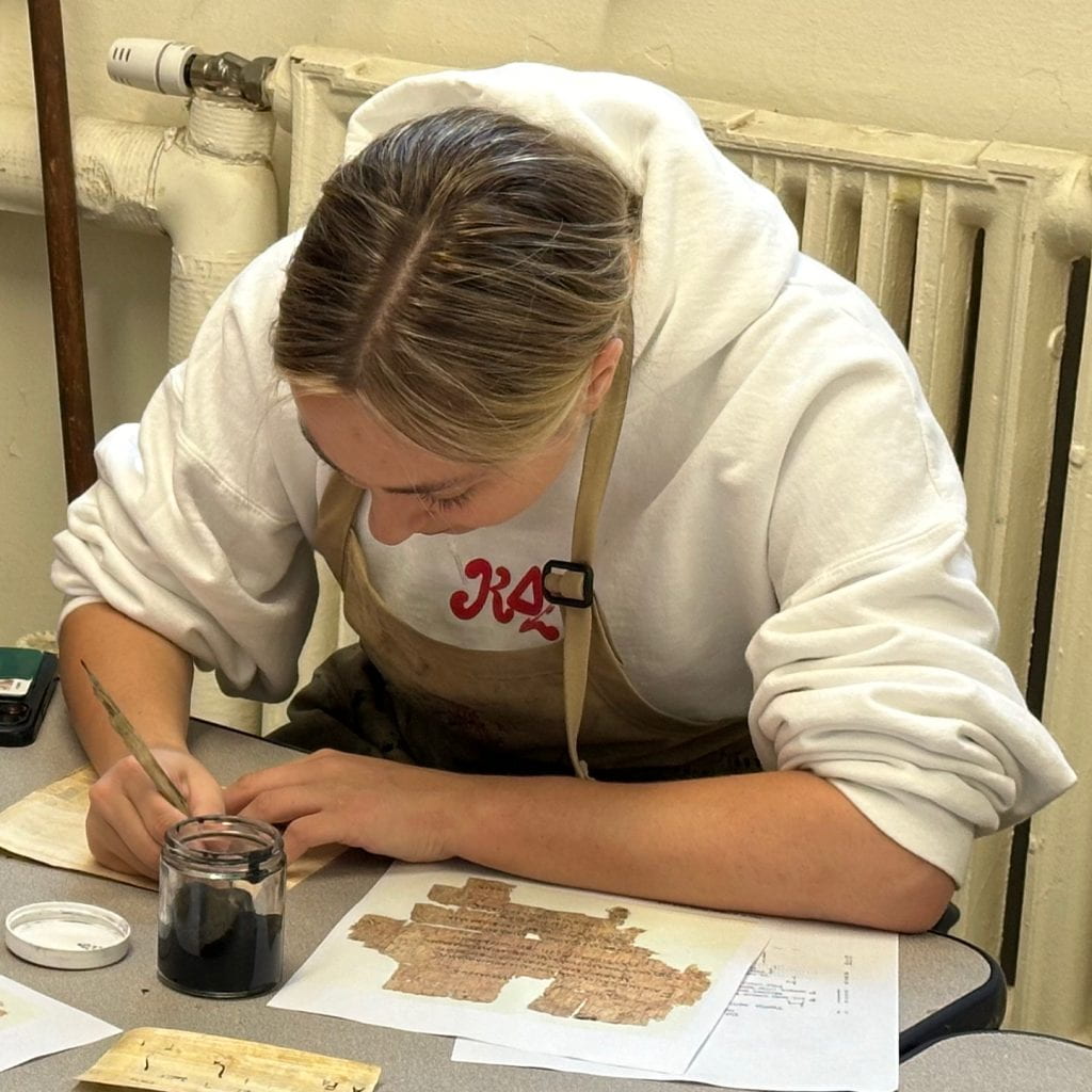 A student in the book arts workshop writes on papyrus using reed pens and lamp black ink