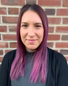 A portrait style photograph of a young white woman with purple hair. She is standing in front of a brick wall.