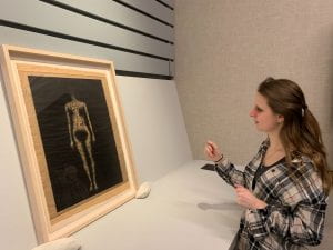 A young woman looking at a framed work of art on paper, that depicts a nude woman, without any features, on a black background.