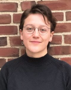 A photograph of a young woman against a brick background. She wears a black turtle neck and glasses.