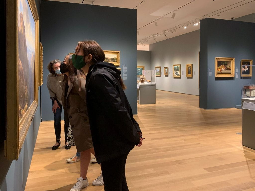 A group of college-aged students on a tour with a museum educator. They are standing close to an American landscape painting, with their hands behind their backs.