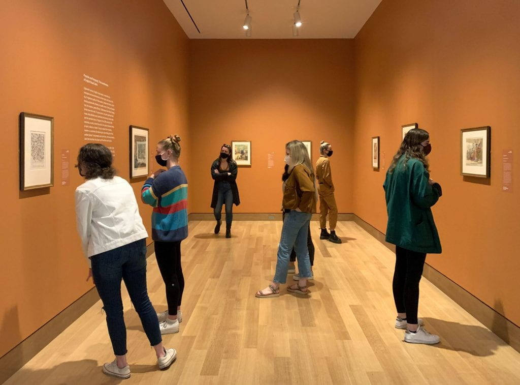A group of college-aged students on a tour, standing in a gallery with orange walls and looking at old English prints.