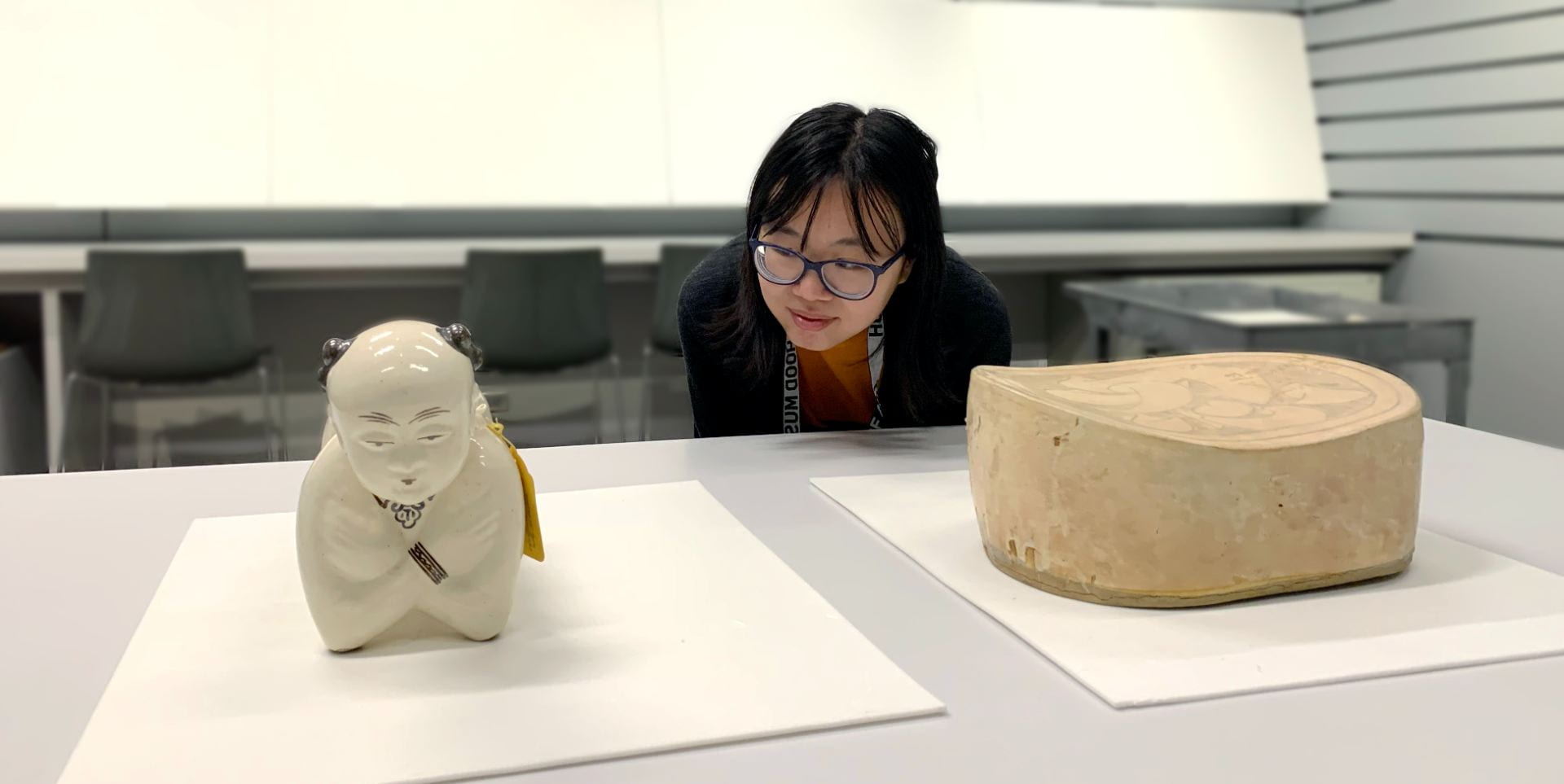A young women leans down to take a closer look at two ancient Chinese ceramic pillows on a table.