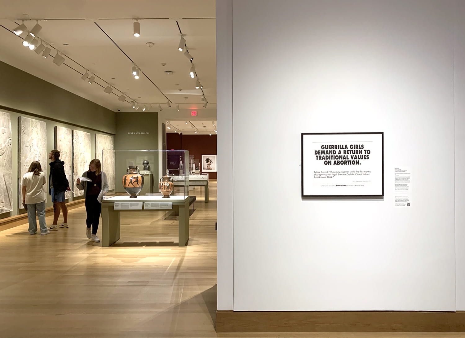A museum gallery with a Guerrilla Girls poster in the foreground, and students explore the galleries beyond.