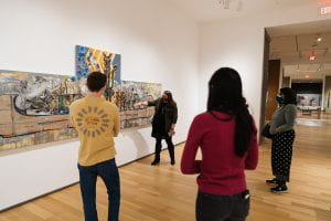A small group of people stand in front of a large painting made of three canvas panels. It depicts a Indigenous American canoe with symbolism piled inside. A curator is speaking to the group of people about the work.