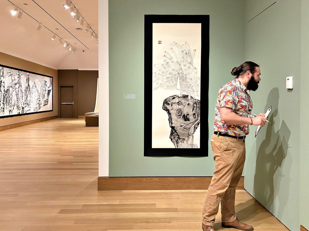 A young man stands in a museum gallery with greens walls and black and white ink drawings on paper. He is checking the humidity of the gallery on a wall thermostat.