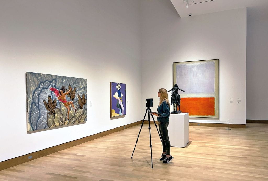 A woman stands with a camera on a tripod in the middle of a museum gallery with heigh ceilings, white walls, and large paintings on the walls. 