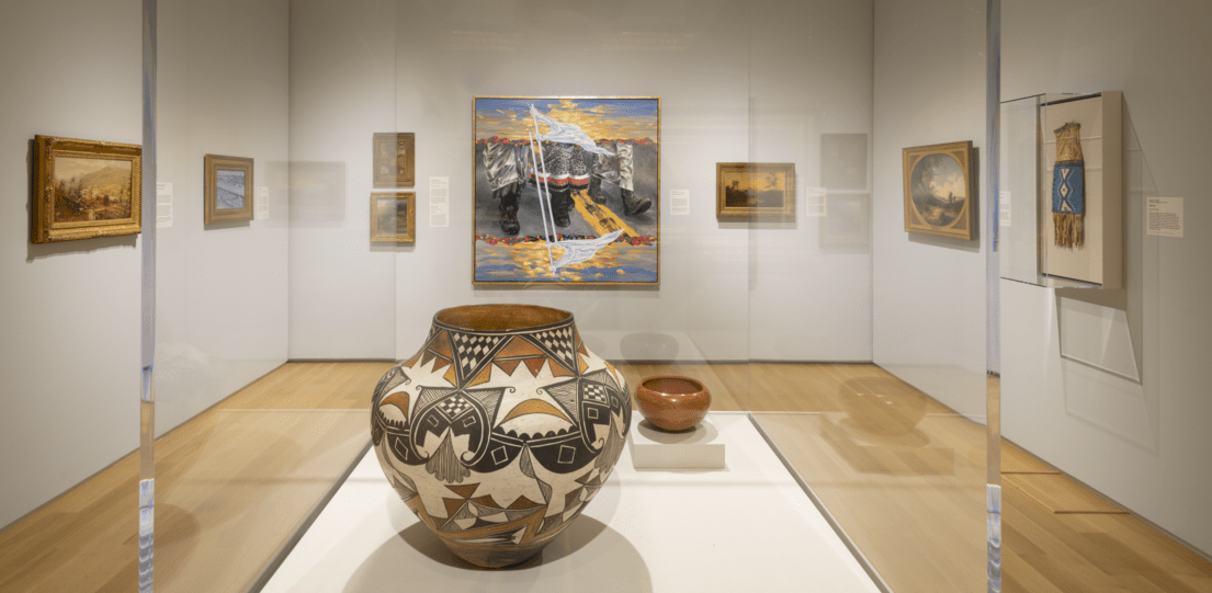 The "Olla Water Jug" sits in a glass case. The glass case sits in a gallery filled with art on the walls.