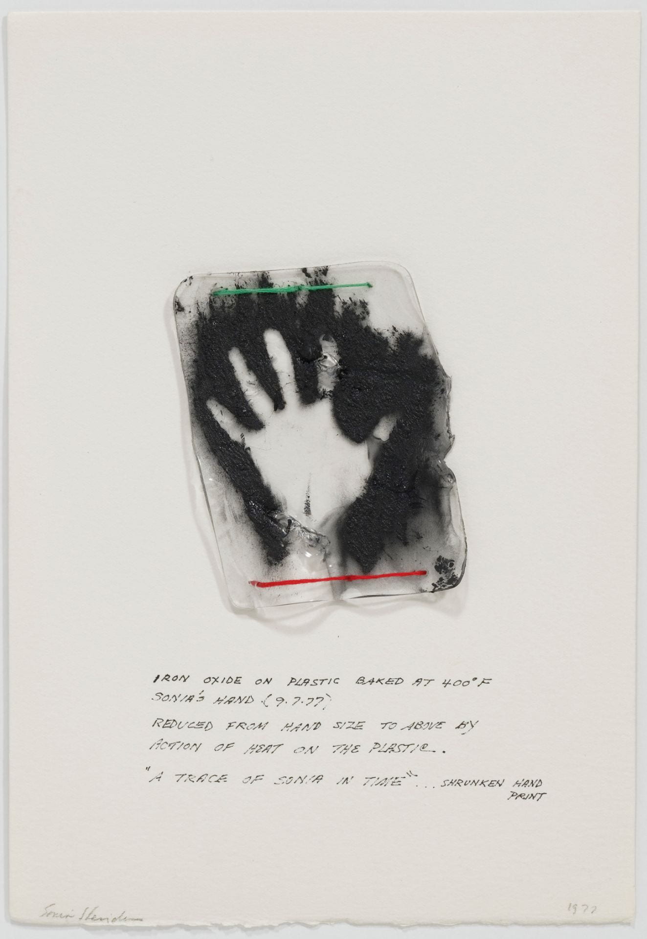 At the center of a white page, the artist has affixed a shrunken plastic replica of her handprint using strands of green and red thread. The handprint appears as a negative, visible as a white print in a cloudlike background of black pigment. Below the fastened object, the artist has inscribed in ink, “IRON OXIDE ON PLASTIC BAKED AT 400 [degree symbol] F / SONIA’S HAND (97.77) / REDUCED FROM HAND SIZE TO ABOVE BY / ACTION OF HEAT ON THE PLASTIC. / ‘A TRACE OF SONIA IN TIME’ . . . SHRUNKEN HAND PRINT.”
