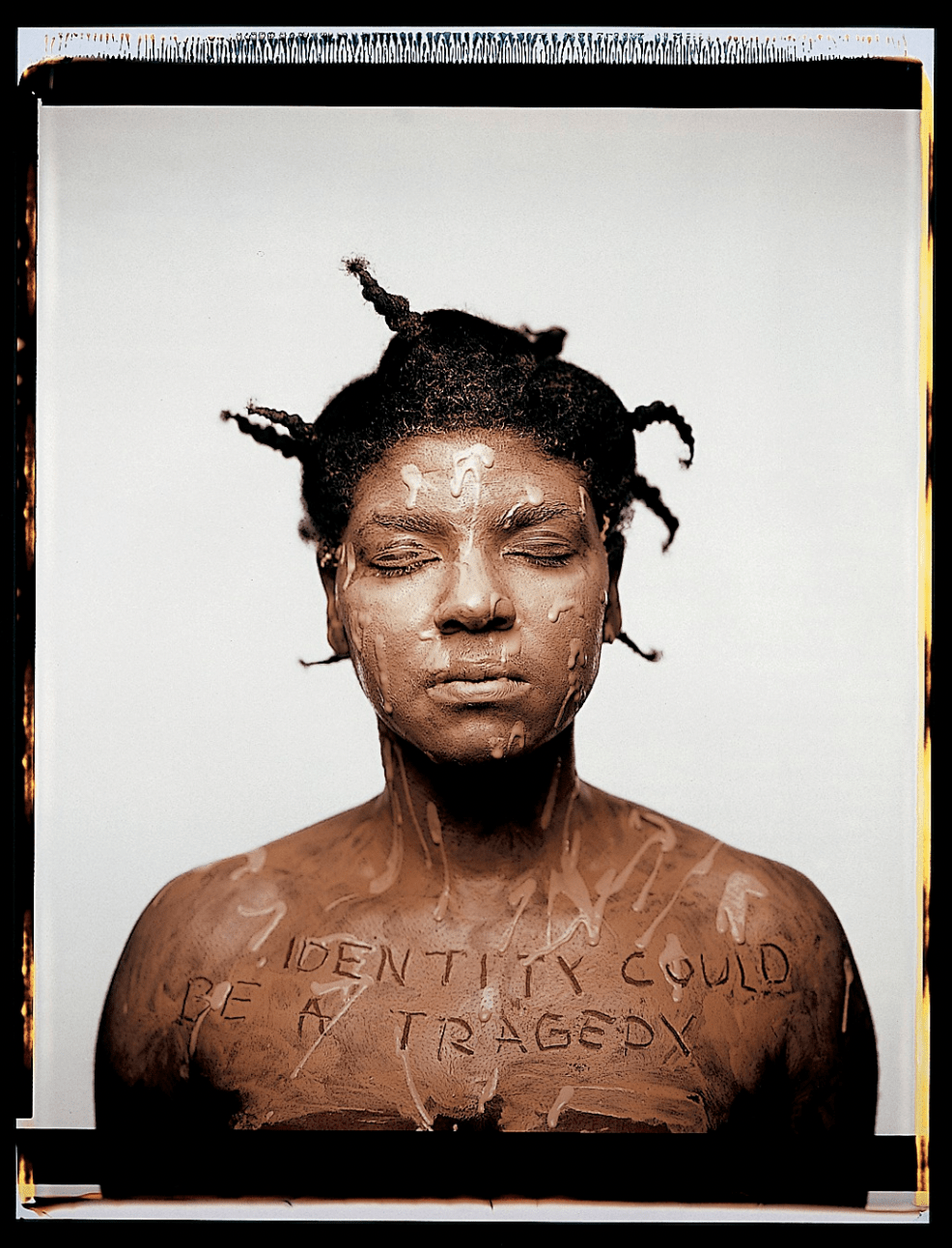 A Polaroid photograph depicts the artist from the shoulders up as she faces the camera with her eyes closed, her bare skin seemingly covered with splattered layers of brown paint. Standing before a spare white background, the artist has inscribed across her chest in capital letters, “IDENTITY COULD BE A TRAGEDY.” 