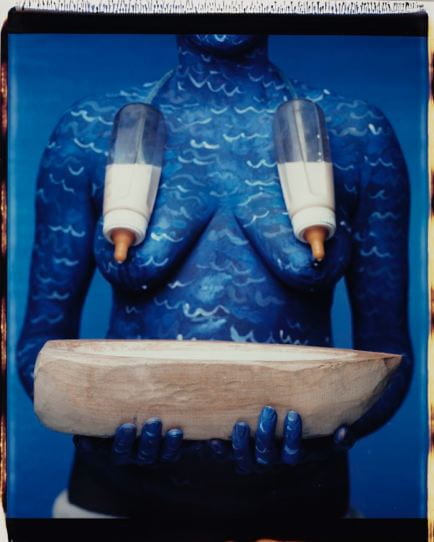 A Polaroid photograph captures the nude torso of the artist covered in deep blue paint accented with white, wavelike lines. Standing in front of a blue background matching the color of her body paint, Campos-Pons has two milk-filled baby bottles strung around her neck, covering her breasts and dripping onto a wooden vessel shaped like a boat, which she holds in both hands in front of her stomach.
